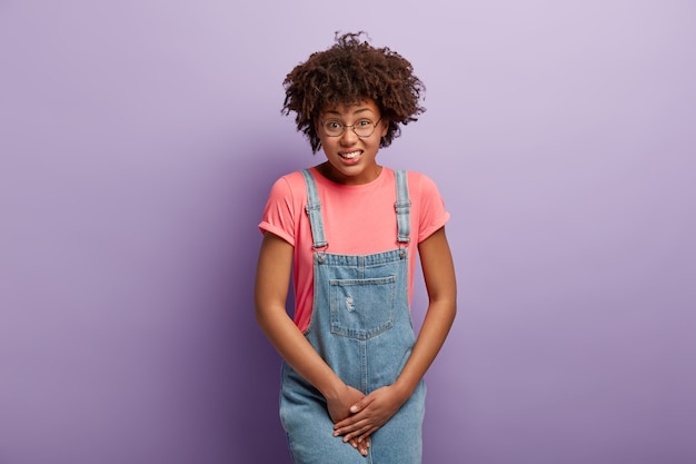 Foto gratuita la donna infelice dalla pelle scura tiene il cavallo, ha bisogno di servizi igienici, ha una situazione problematica, indossa una maglietta rosa e jeans sarafan, soffre di cistite, isolata su un muro viola. persone e urgenza