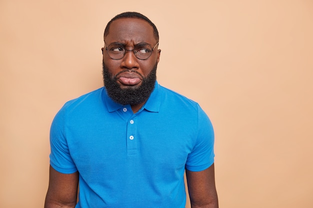 Unhappy discontent man has sulking expression wants to cry because of problems purses lips looks away wears big spectacles basic casual blue t shirt isolated over brown wall