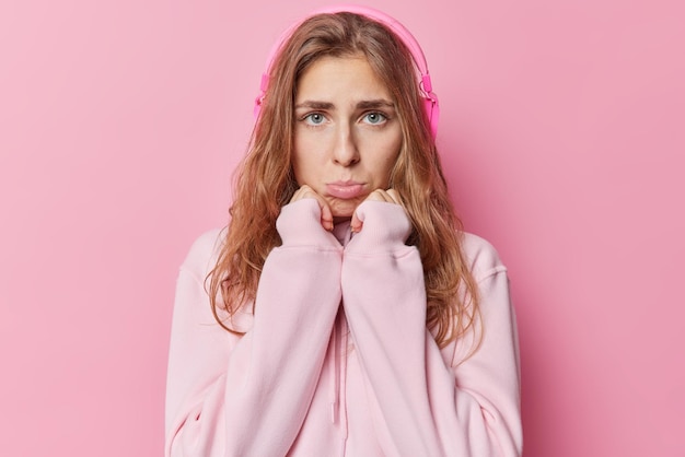 Free photo unhappy displeased beautiful woman with blue eyes purses lis keeps hands under chin feels frustrated has bad mood listens music via headphones wears casual hoodie isolated over pink background