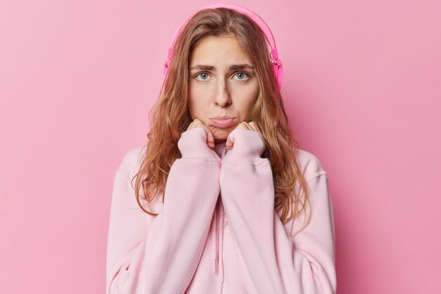 Unhappy displeased beautiful woman with blue eyes purses lis keeps hands under chin feels frustrated has bad mood listens music via headphones wears casual hoodie isolated over pink background