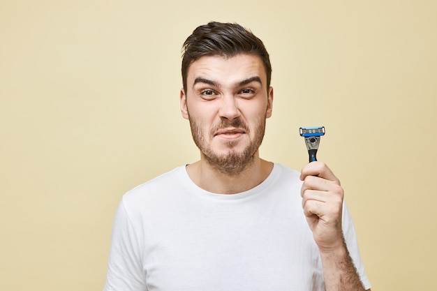 unhappy displeased young brunette man with bristle grimacing does not want to shave his beard, hating shaving process, having sensitive skin, posing isolated with razor in hands