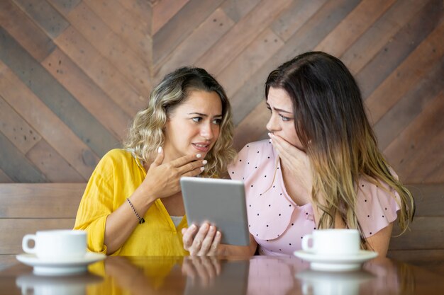 Unhappy female friends reading shocking news on tablet computer