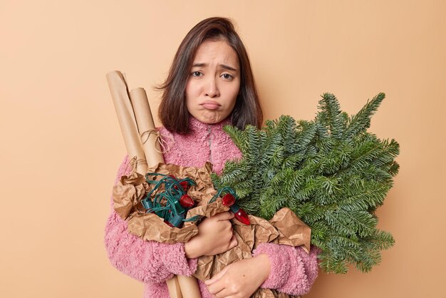 Unhappy gloomy brunette woman with eastern appearance holds holiday items for decoration prepares for New Year celebration wears winter coat isolated over beige background. Christmas Eve concept