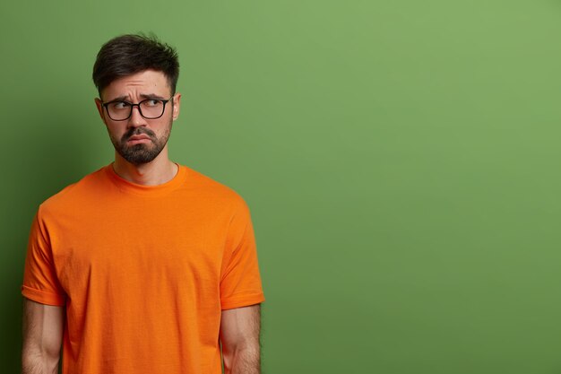 Unhappy gloomy young European man looks upset and disappointed, wears casual orange t shirt and spectacles, feels uneasy and moody, stands against green wall, copy space for your promotion.