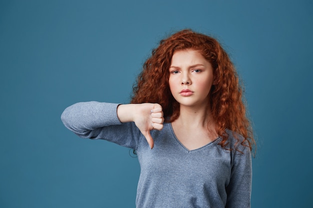 Foto gratuita infelice bella donna con i capelli ricci rossi e le lentiggini con espressione triste e stanca, mostrando il pollice verso il basso.