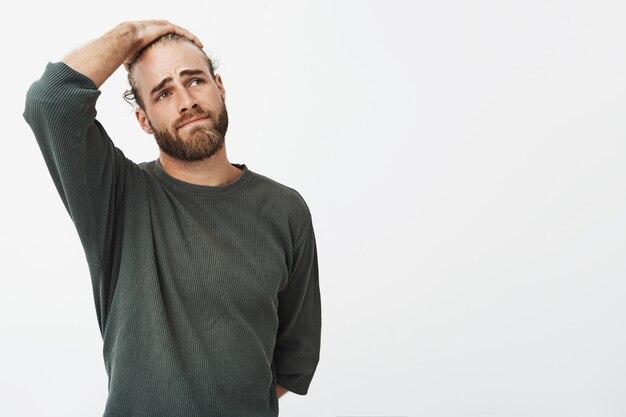 Unhappy good-looking young man with fashionable hairstyle and beard holding head with hand