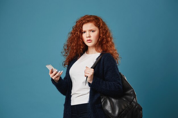 Unhappy handsome ginger student woman with freckles holding phone with nervous look, after talk with teacher about exam marks.