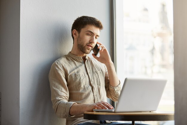 暗い髪の不幸なハンサムな男、カフェに座って、ラップトップコンピューターで作業し、電話で不満の顧客と話しています。