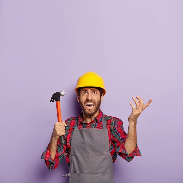 Free photo unhappy handyman holds hammer, focused above with annoyance, repairs something with building tool in workshop, wears hardhat, shirt and apron. foreman inspector at work, does repairing alone