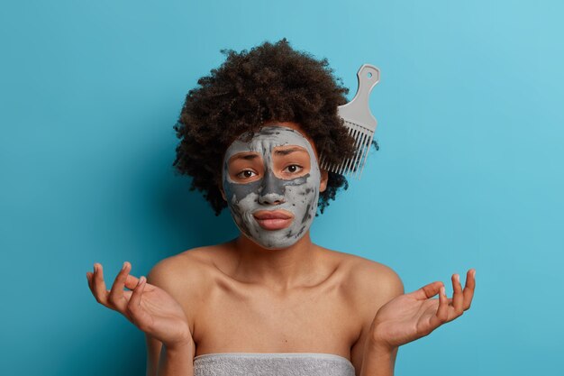 不幸な躊躇しているアフリカ系アメリカ人の女性は、くしが縮れた自然な髪に刺さっていて、困惑して手を横に広げ、くしの仕方がわからず、化粧品のフェイスマスクを着用し、体の周りにバスタオルを着用しています