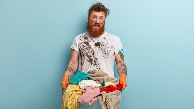 太い毛の不幸な憤慨した赤毛の男、だらしのないTシャツ、ゴム手袋を着用し、洗濯物の山を運び、汚れた顔をして、青い壁に立ち、家で洗濯をしたくない