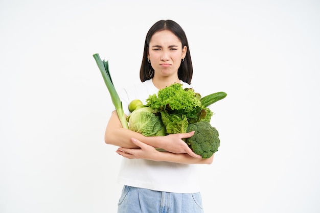 Free photo unhappy korean woman holding vegetables feeling unhappy on diet eating raw green food white backgrou