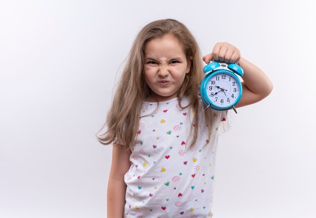Unhappy little school girl wearing white t-shirt holding alarm clock on isolated white wall