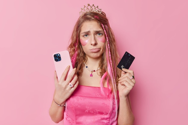 不幸な長い髪の若い女性は携帯電話を持っており、クレジットカードはオンラインで支払いを行いますドレスクラウンとネックレスはパーティーの準備をしますピンクの背景の上に分離された美容パッチを適用します