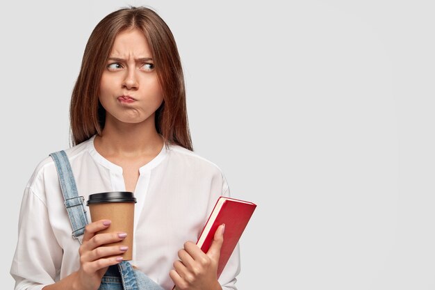 Unhappy lovely female student feels displeased from learning all time, holds cup of takeaway coffee and book, thinks about rest, has sullen expression