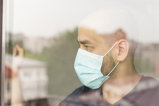 Unhappy man looking through window wearing protection mask during self isolation