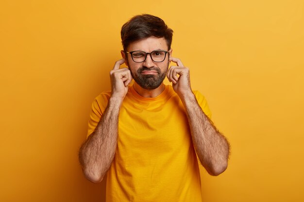 Unhappy man plugs ears with fingers for not hear anymore, ignores loud music coming from neighbours, feels discomfort, stands against yellow wall, disturbed by annoying sound or noise.
