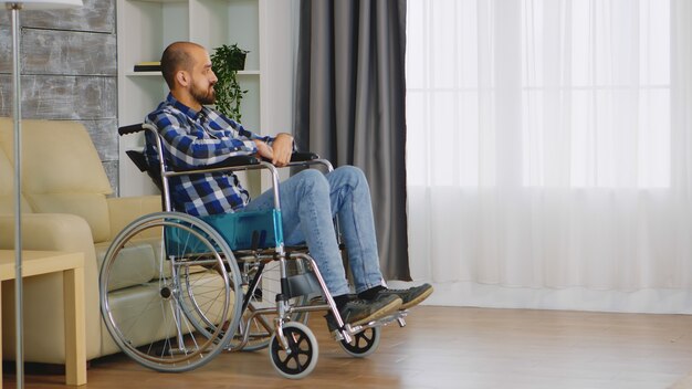 Несчастный человек в инвалидной коляске в гостиной, глядя в окно.