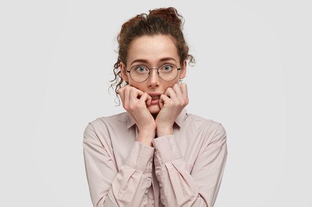 Unhappy nervous young female with worried expression, bites finger nails, looks anxiously directly, wears spectacles, dressed in fashionable clothes, stands against white wall.