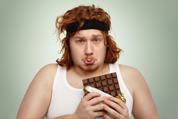 Unhappy obese redhead man wearing black sports headband and white tank top having sad and frustrated look