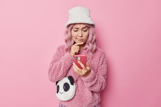 Unhappy pink haired young Asian woman looks seriously at smartphone screen keeps hand on chin has thoughtful expression wears winter panama and coat carries bag isolated over rosy background