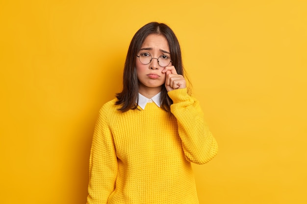 Unhappy sad dejected woman with eastern appearance rubs tears wants to cry feels desperate has problems in life wears round spectacles and casual jumper