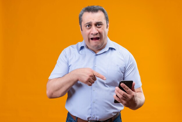 青い縦縞のシャツを着た不幸な悲しい男がオレンジ色の背景の上に立っている間彼の指を携帯電話に向ける