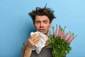 Free photo unhappy sick european man suffers from rhinitis and allergy, sneezes in napkin, has problems with breathing, holds blooming plant, looks frustrated , poses over blue wall, feels unwell