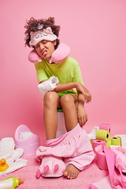 Несчастная сонная женщина позирует на унитазе, страдает от запоров, носит подушку для шеи с завязанными глазами и футболку, окруженную множеством вещей в ванной, изолированных на розовом фоне.