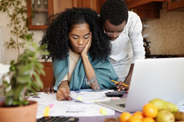 Несчастная и напряженная молодая африканская женщина сидит за кухонным столом с бумагами и ноутбуком, пытаясь сократить количество домашних расходов, одновременно составляя семейный бюджет вместе со своим мужем
