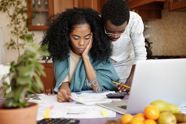 Unhappy and stressed young African woman sitting at kitchen table with papers and notebook computer, trying to cut amount of domestic expenses while doing family budget together with her husband