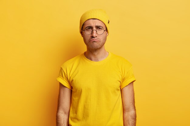 Несчастный, неудовлетворенный парень ухмыляется, имеет мрачное выражение, носит желтую шляпу и футболку.