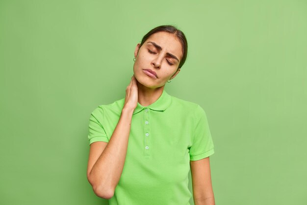 Unhappy woman feels pain in neck grimaces from pain suffers without painkillers has problems with health keeps eyes closed tilts head dressed in casual t shirt isolated over green wall