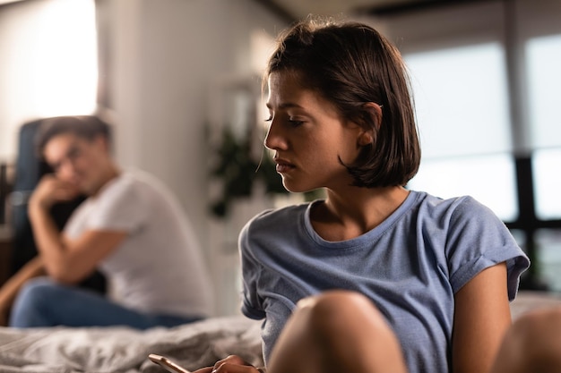 Foto gratuita donna infelice che manda un sms sul cellulare dopo una discussione con il suo ragazzo a casa l'uomo è sullo sfondo