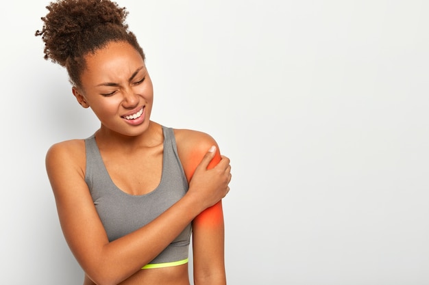 Free photo unhappy woman with curly combed hair, has painful feelings in left shoulder, clenches teeth, wears casual grey sport bra