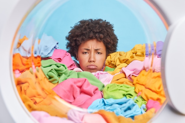 Unhappy woman with curly hair has tired expression does laundry at home drowned in multicolored clothes shows only head feels discontent