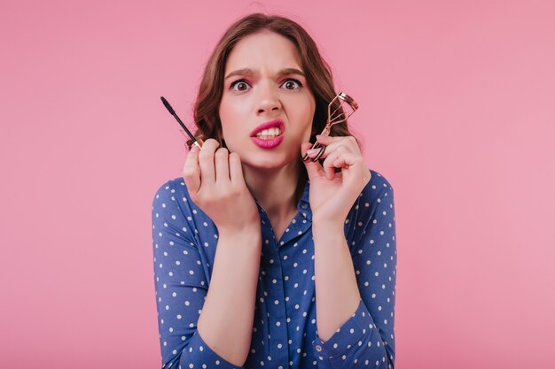 Unhappy woman with wavy hair doing her makeup before date. Nervous girl in blue attire curls eyelashes on pink wall.