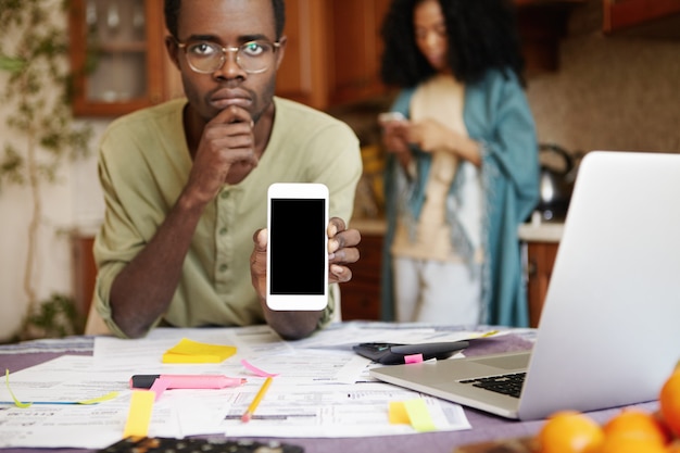 Несчастный молодой африканский мужчина в очках сидит за столом с бумагами, ноутбуком и калькулятором, подсчитывая семейный бюджет, держа в руке сотовый телефон