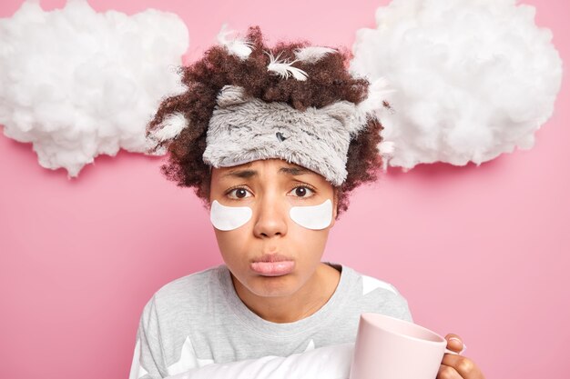 不幸な若いアフロアメリカ人女性は悲しいことにカメラを見てパッチを適用しますsleepmaskパジャマは上のピンクの壁の白い雲の上に孤立して目覚めた後にコーヒーを飲みます