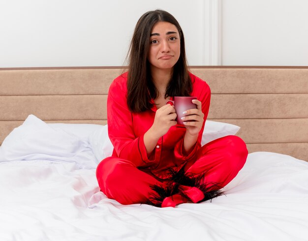 Unhappy young beautiful woman in red pajamas sitting on bed with cup of coffee looking at camera with sad expression in bedroom interior on light background