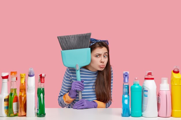 Unhappy young cleaning service employee holds broom, uses numerous cleaning substances, arent ready to start working, has negative attitude to work