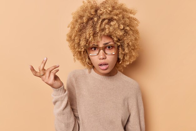 Unhappy young curly haired woman feels indignant keeps hand raised feels clueless dressed in casual jumper isolated over beige studio background cannot understand something So what did you say