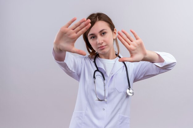 Unhappy young female doctor wearing medical robe and stethoscope doing stop gesture  on isolated white wall with copy space