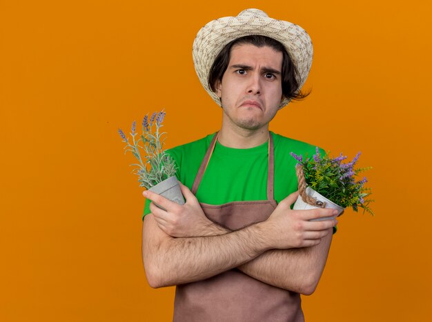 オレンジ色の背景の上に立っている悲しい表情でカメラを見てうんざりした植物を保持しているエプロンと帽子の不幸な若い庭師の男