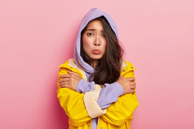 Unhappy young girl feels lonely and cold, looks sadly at camera, keeps hands crossed, wears hoody and raincoat, being dejected by bad weather