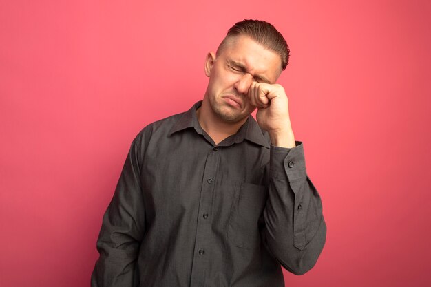 Unhappy young handsome man in grey shirt rubbing his eyes crying standing over pink wall