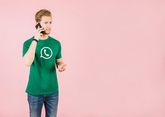 Unhappy young man talking on cellphone against pink background