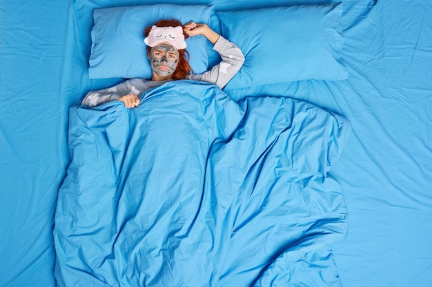 불행한 젊은 여성이 기분이 좋지 않아 슬프게 보이며 파란색 담요 아래 침대에 누워 얼굴에 영양 미용 마스크를 착용합니다.