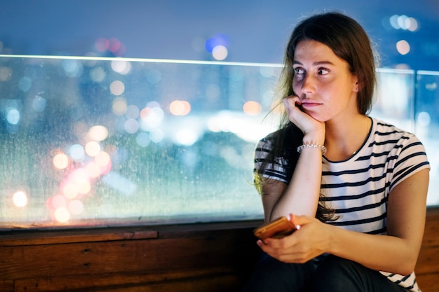Foto gratuita giovane donna infelice che tiene uno smartphone nel paesaggio urbano di sera