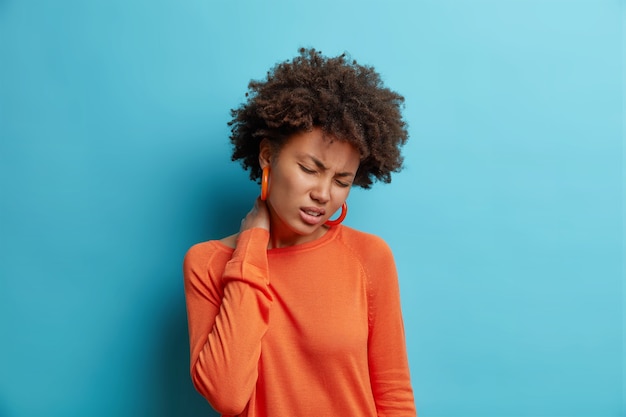 Unhappy young woman suffers from pain neck feels tired massages neck feels discomfort closes eyes wears casual orange jumper isolated over blue wall
